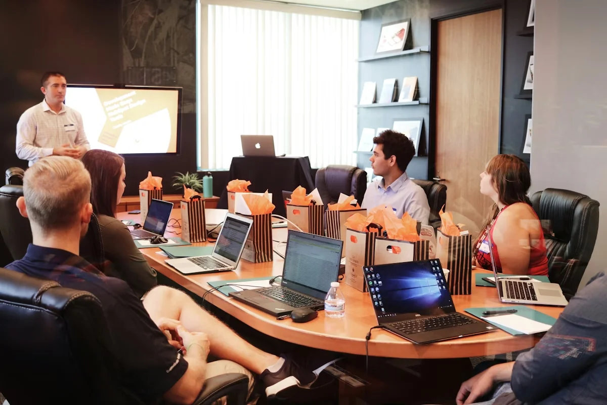 Consultant presenting digital strategy to a team around a conference table — five people with laptops facing the screen